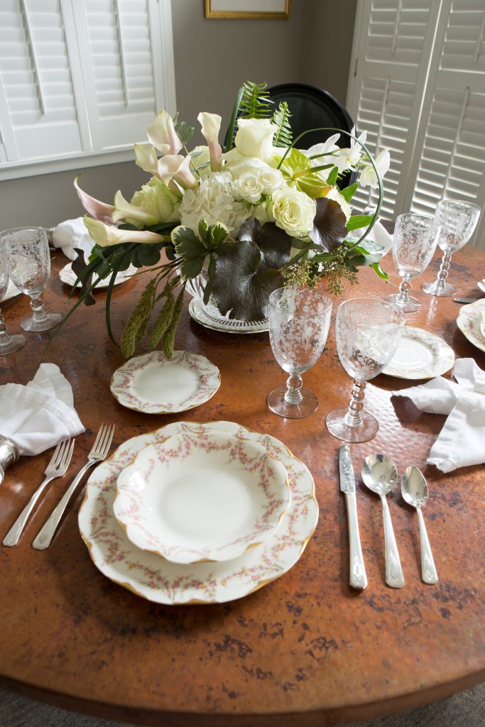  Shari’s table is set with china, crystal, and flatware from her mother, Lynette Hutchinson, that previously belonged to her grandmother, Pretto Zeigler. The spectacular flower arrangement by Corky Robinson at The Blossom Shop contains white hydrangeas, green garden roses, white moth orchids, blush and green callas, and green anthurium. 

