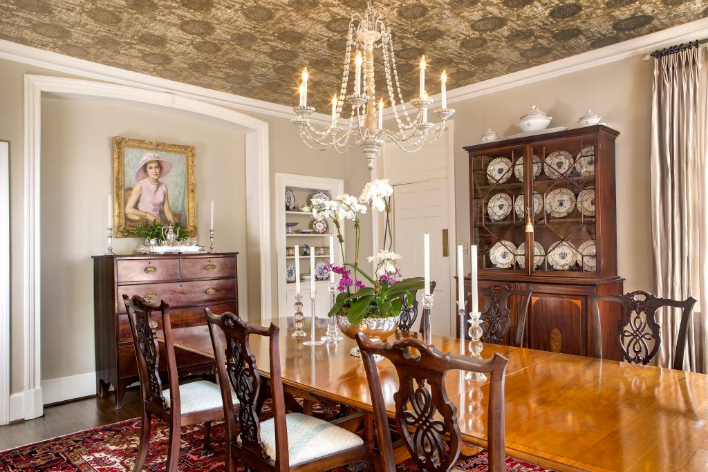 Previous owner of the Adger home Lilla Hoefer wallpapered the ceiling of the dining room with a gold medallion patterned paper, which Lura Folline kept as a beautiful accent for her family antiques. A portrait of Lura’s maternal grandmother, Rosalind Vareen Lanier, hangs above the central chest. 