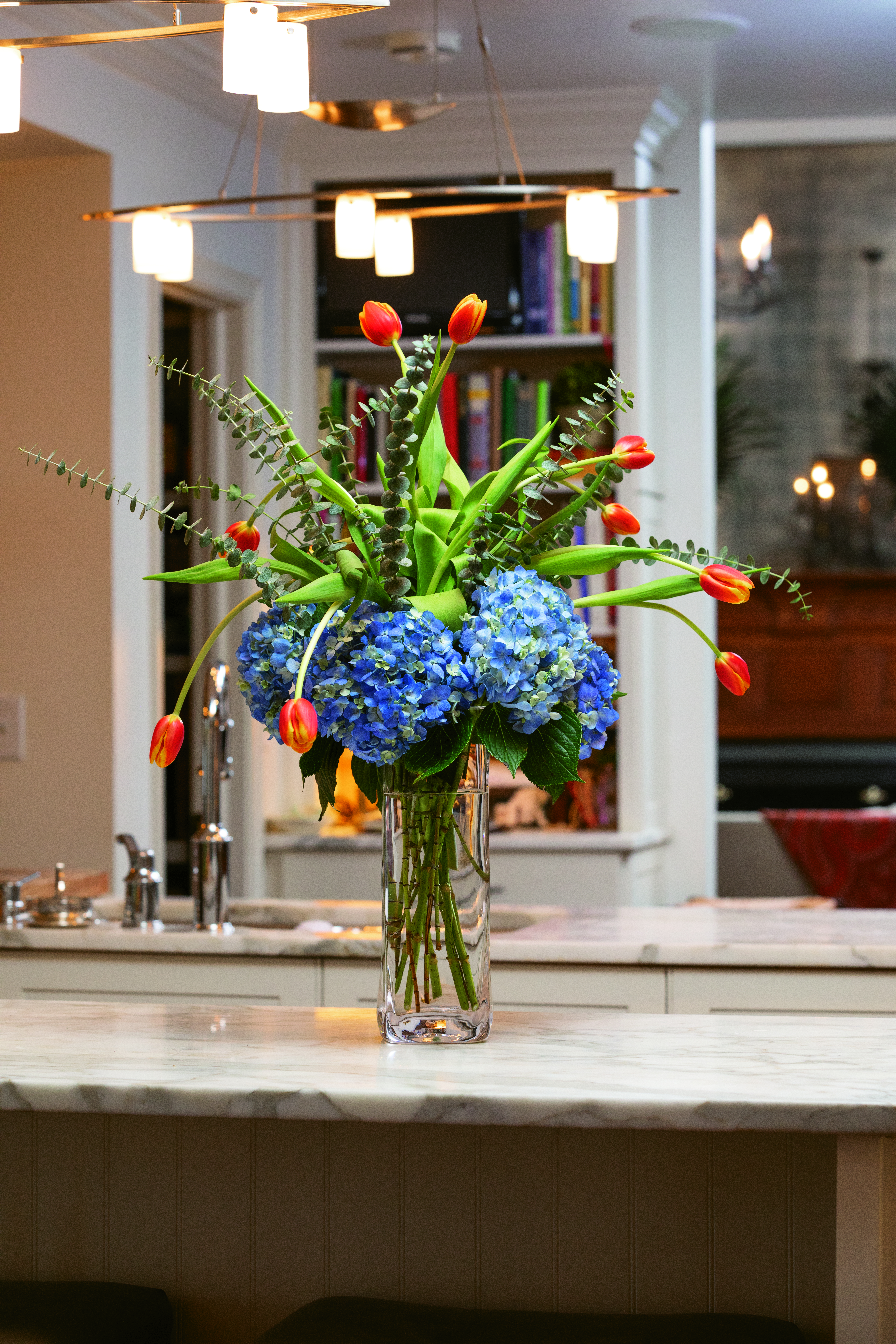 To build this arrangement, place large blue hydrangea blooms at the base of a tall cylinder vase, then artfully insert strong, tall eucalyptus in the center of the arrangement followed by tulips to add a touch of whimsy. 