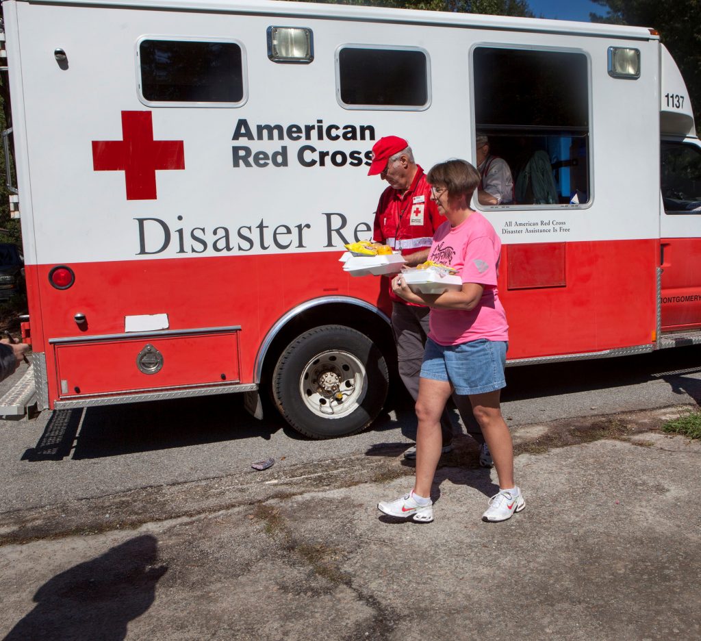 The Red Cross provides hot meals, snacks, and water to residents affected by South Carolina flooding. Red Cross volunteer Roger Blinn helps a local resident carry a meal and water to her home, October 2015. Photography courtesy of the American Red Cross