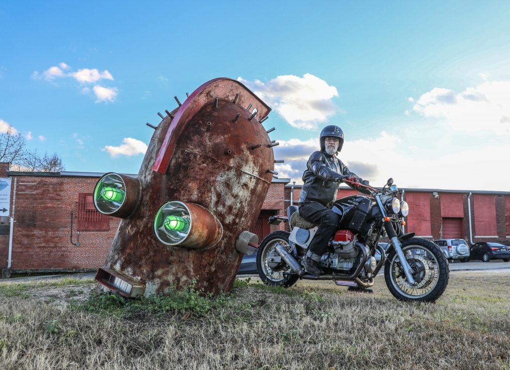 Clark, astride his Moto Guzzi, poses beside one of his robot creations outside his Huger Street studio.  