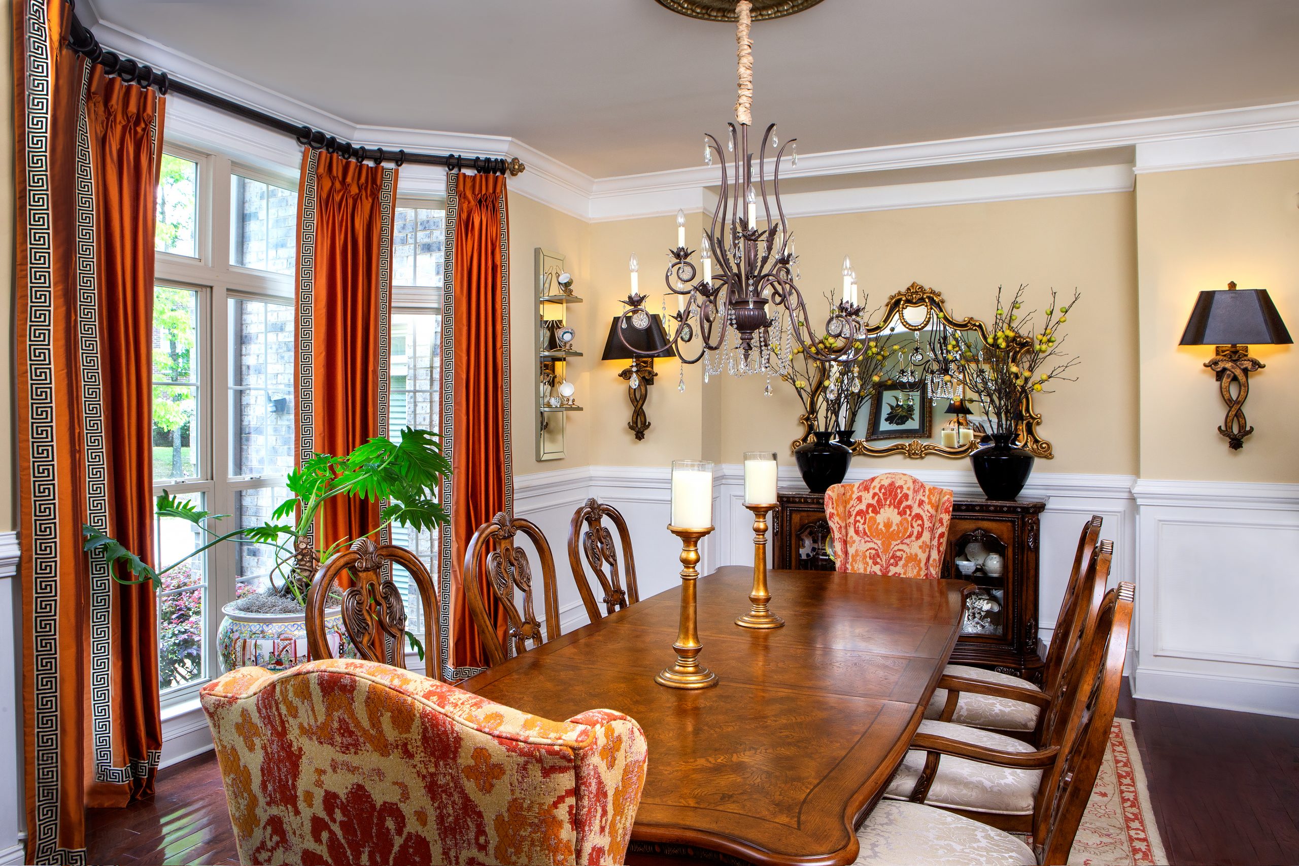 The large dining room has a fabulous ambiance with warm yellow walls and a gracious bay window. Host and hostess chairs in ginger and orange on cream complement the rug, and a handsome mirror over the buffet reflects the entrance hall and sparkling chandelier.