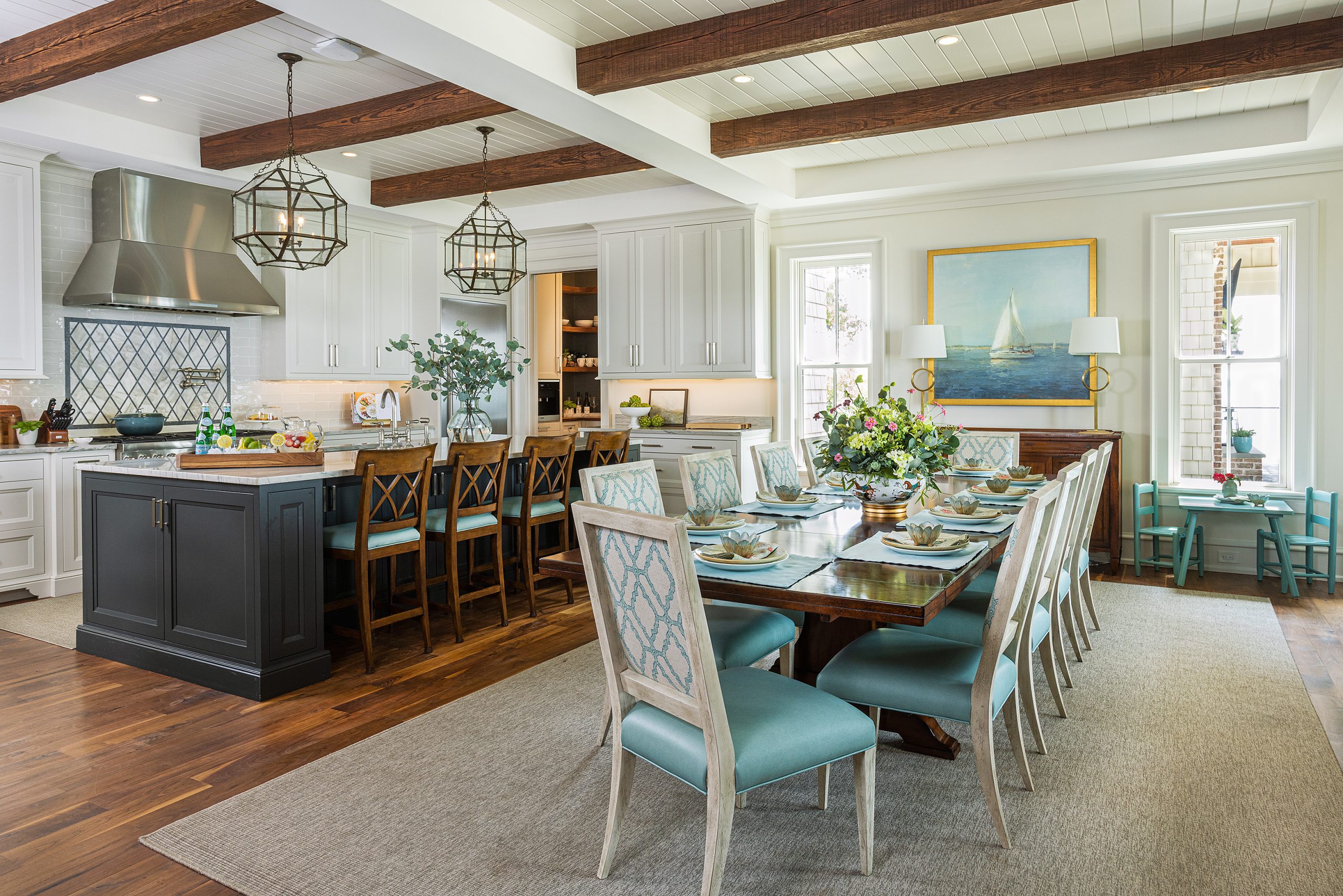 The combined kitchen and dining room is where the family spends most of their time. The aqua color palette throughout the home reflects the water. When Amy prepares food at her island, she has a magnificent view of the sunset. Flowers courtesy of Amy’s sister, Allison Plumblee. Home styled for photography by Sheila Andrade with Black Barn Home
