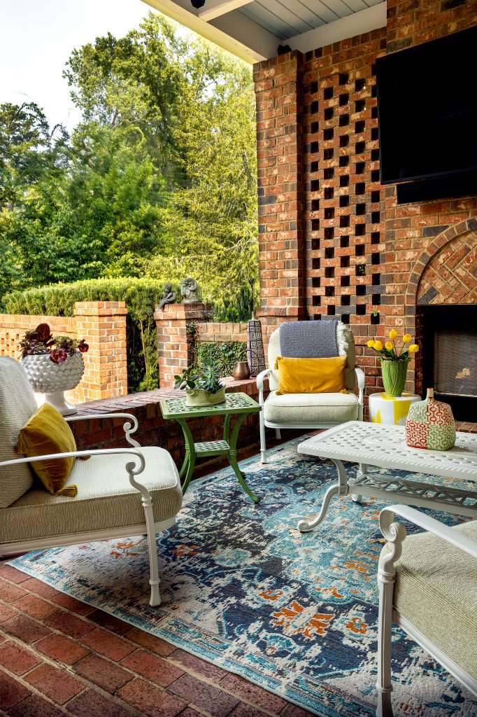 The wide, brick back porch is well used and enjoyed. A conversation space with a sofa and two chairs surrounding a coffee table is opposite a gas burning fireplace under a television and a chair foursome. Matching rugs for both seating spaces give the porch a cohesive look. A few steps down is a partially enclosed brick patio with flower beds, bird feeders, a fountain, and black wrought iron seating. 

