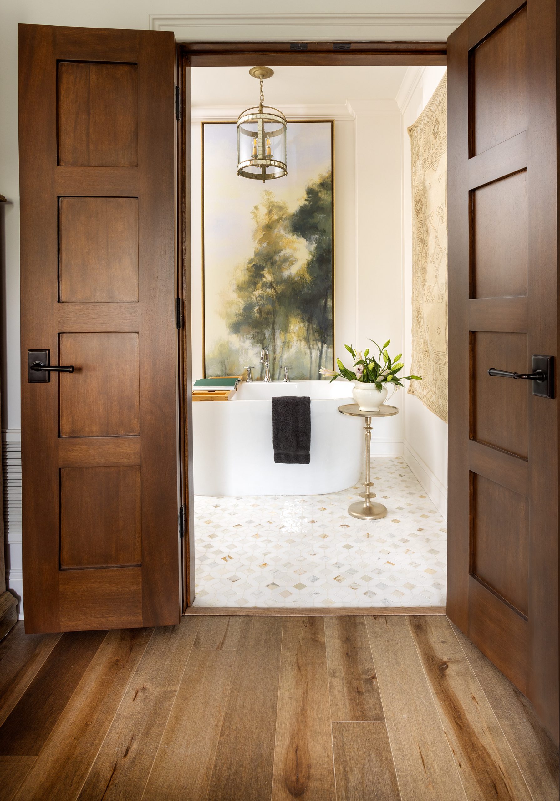 Dark mahogany double doors connect the primary bedroom suite to the bath, opening to a soaking tub that perches beneath a tall abstract painting.
